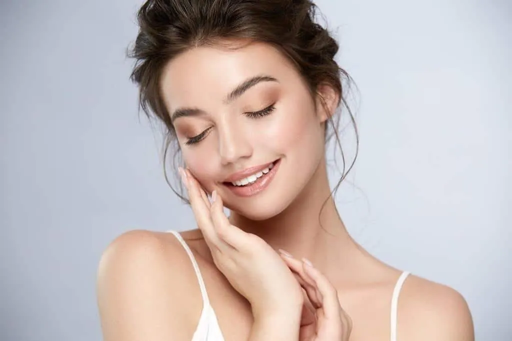 Žena s úsměvem si jemně přidržuje tvář, má přirozený make-up.