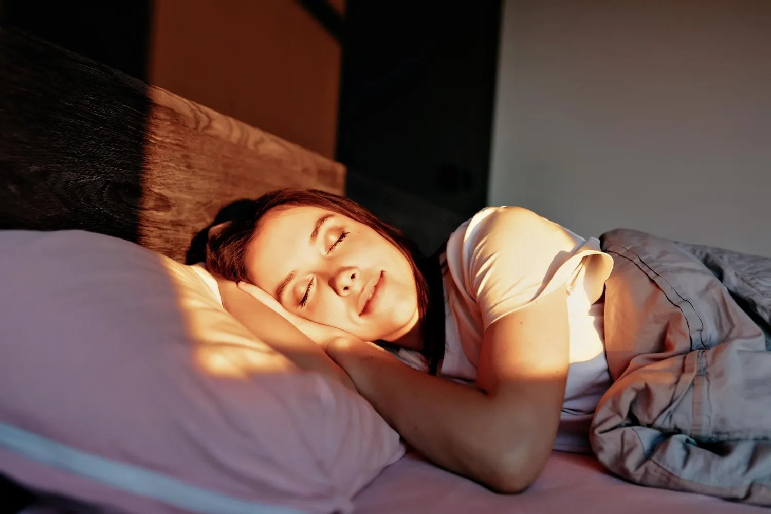 Žena spí na posteli s růžovými polštáři, sluneční světlo osvětluje její tvář.