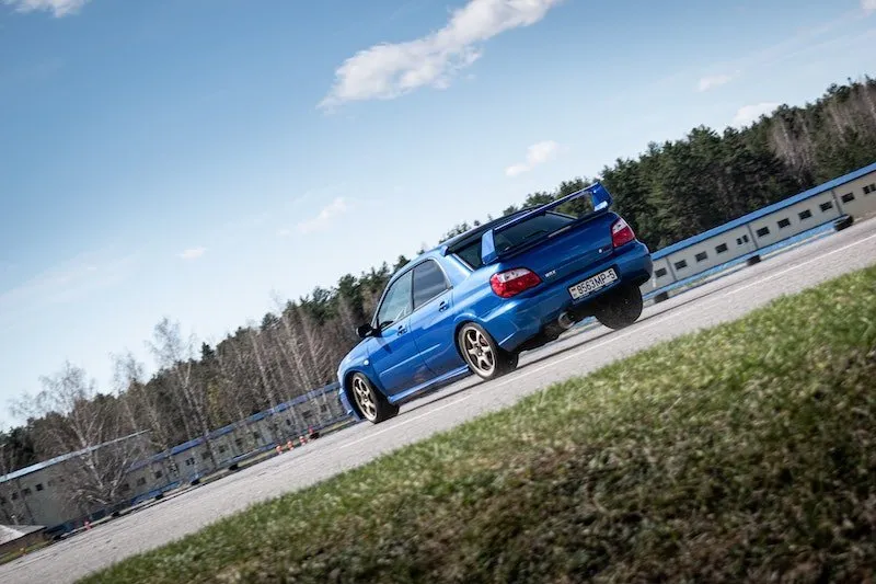 Modrý Subaru s aerodynamicým zadním spoilerem na silnici, lesní pozadí.