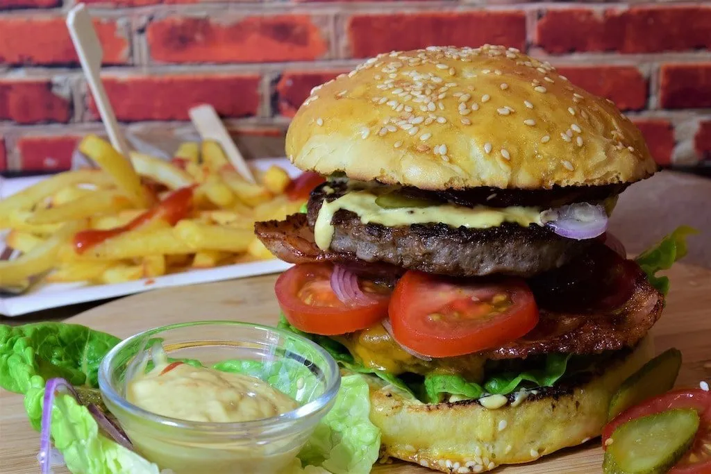 Šťavnatý hamburger se slaninou, zeleninou a omáčkou, vedle hranolky.