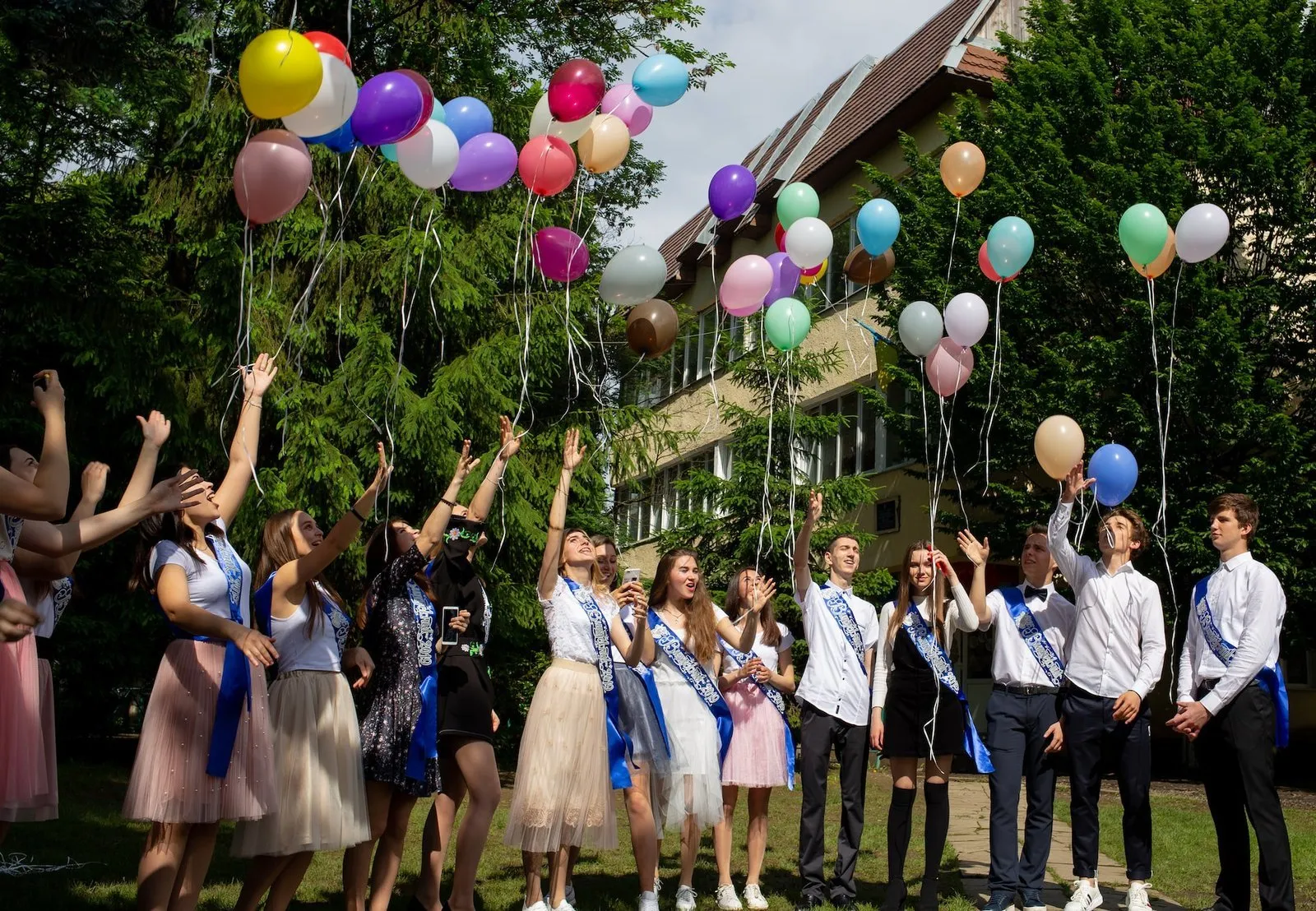 Skupina maturantů v bílých košilích s barevnými balónky a šerpami.